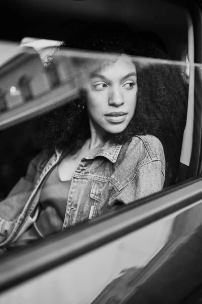 Potret hitam dan putih dengan refleksi dari wanita cantik Afrika Amerika sopir berambut hitam duduk di mobil baru, menikmati perjalanan. Mengemudi kursus dan konsep asuransi jiwa — Stok Foto