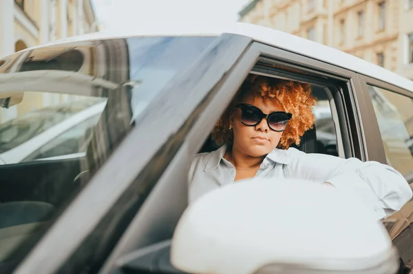 หญิงสาวชาวแอฟริกันอเมริกันที่มีความสุข ผู้ขับขี่ผมแดงในแว่นตากันแดดนั่งอยู่ในรถสีน้ําตาลใหม่ยิ้มมองกล้องเพลิดเพลินกับการเดินทาง หลักสูตรการขับขี่และแนวคิดประกันชีวิต — ภาพถ่ายสต็อก