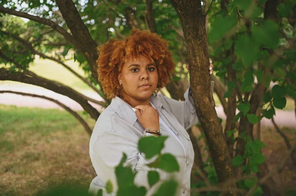 Potret mode wanita muda cantik secara alami ditambah ukuran wanita Afrika Amerika dengan pakaian celana panjang terang dengan rambut merah afro yang berpose di taman alam dalam dedaunan hijau. Perbedaan dan konsep positif tubuh. — Stok Foto