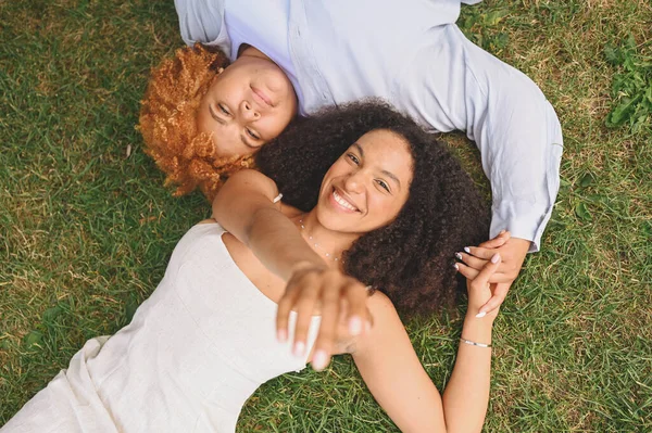 Pasangan muda Afrika-Amerika lesbian yang bahagia berbaring di rumput sambil tertawa sambil menjulurkan tangan sambil menutup kamera, pemandangan puncak musim panas. Konsep komunitas LGBT. Perempuan teman menikmati saat-saat cinta. — Stok Foto
