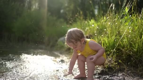 Lilla roliga söta blonda flicka barn småbarn i gul våt body suit spelar vid sjön stranden vid solnedgången utanför. Baby med lera på händer och ben. Vattenaktiviteter på sommaren. — Stockvideo