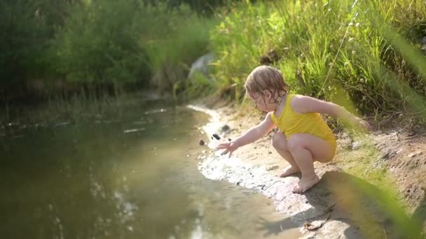 Kis vicces aranyos szőke lány gyermek csecsemő sárga nedves testruha játszik a tó partján naplementekor kívül. Baba sárral a kezén és a lábán. Vízügyi tevékenységek nyáron. — Stock videók