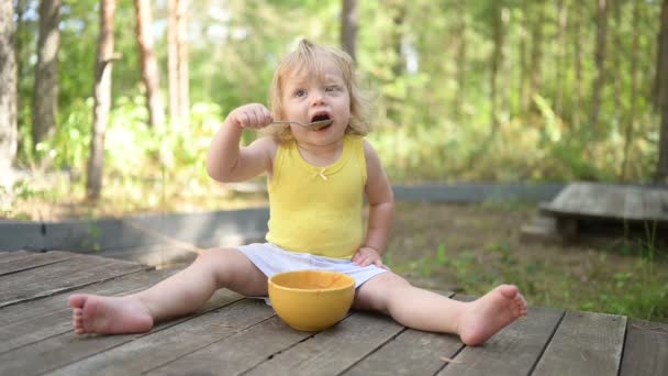 더러운 옷을 입고 어린 금발의 귀여운 여자 아이가 여름에 밖에서 노란 접시에서 숟가락으로 아기 음식이나 야채 퍼리를 먹는 것을 볼 수있다. 건강 한 어린 시절의 개념. — 비디오