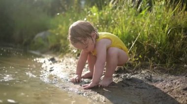 Dışarıda, göl kıyısında gün batımında oynayan sarı tulumlu komik sarışın kız çocuğu. Ellerinde ve bacaklarında çamur olan bebek. Yazın su aktiviteleri.