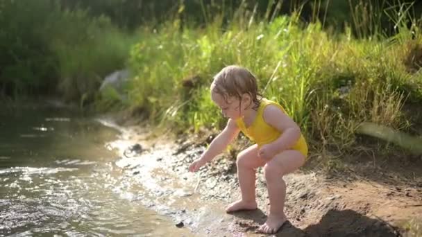 Mała zabawna blondynka dziecko maluch w żółtym mokrym body gra nad brzegiem jeziora o zachodzie słońca na zewnątrz. Dziecko z błotem na rękach i nogach. Aktywność wodna w lecie. — Wideo stockowe