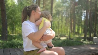 Annesinin kollarında bukleleri olan küçük tatlı bebek sarışın kız. Anne ve kızı yazın kırsal alanda ön bahçede birlikte vakit geçiriyorlar. Sağlıklı mutlu aile çocukluğu kavramı.