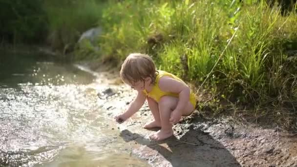 Piccola ragazza bionda simpatica divertente bambino in tuta gialla bagnata che gioca sulla riva del lago al tramonto all'esterno. Bambino con fango su mani e gambe. Attività acquatiche in estate. — Video Stock