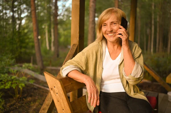 Teknologi, konsep orang tua - wanita tua yang tersenyum bahagia berbicara menggunakan ponsel di luar rumah di teras kayu di taman musim panas di halaman belakang desa — Stok Foto