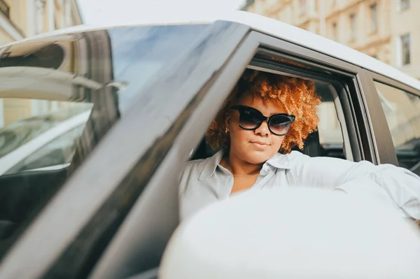หญิงสาวชาวแอฟริกันอเมริกันที่มีความสุข ผู้ขับขี่ผมแดงในแว่นตากันแดดนั่งอยู่ในรถสีน้ําตาลใหม่ยิ้มมองกล้องเพลิดเพลินกับการเดินทาง หลักสูตรการขับขี่และแนวคิดประกันชีวิต — ภาพถ่ายสต็อก