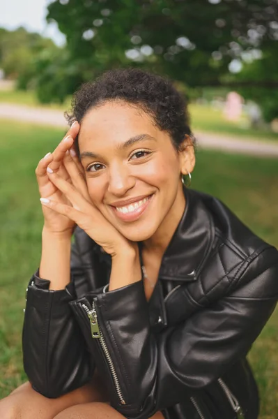 Fashion close-up portret van aantrekkelijke jonge natuurlijke schoonheid Afro-Amerikaanse vrouw met afro haar in zwart lederen jas poseren in natuurpark in groen gebladerte. Perfecte tanden en glimlach. — Stockfoto