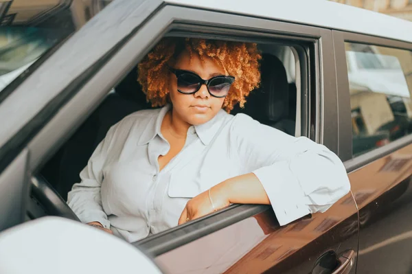 Happy young tersenyum muda Afrika Amerika wanita berambut merah pengemudi kacamata matahari duduk di mobil baru coklat, tersenyum melihat kamera menikmati perjalanan. Mengemudi kursus dan konsep asuransi jiwa — Stok Foto