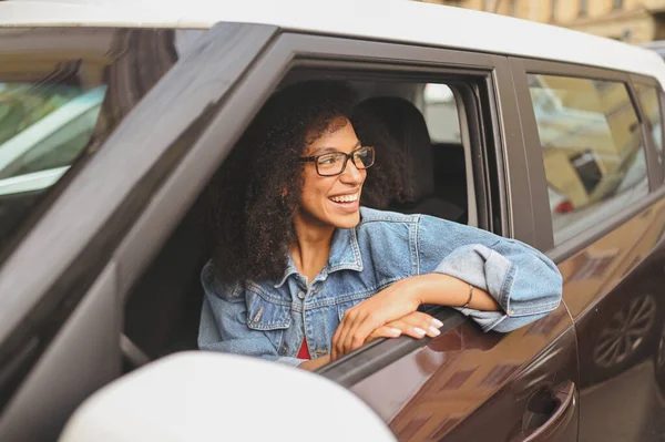 หญิงสาวชาวแอฟริกันอเมริกันที่มีความสุข คนขับรถผมดําในแว่นตานั่งอยู่ในรถสีน้ําตาลใหม่ยิ้มมองกล้องเพลิดเพลินกับการเดินทาง หลักสูตรการขับขี่และแนวคิดประกันชีวิต — ภาพถ่ายสต็อก