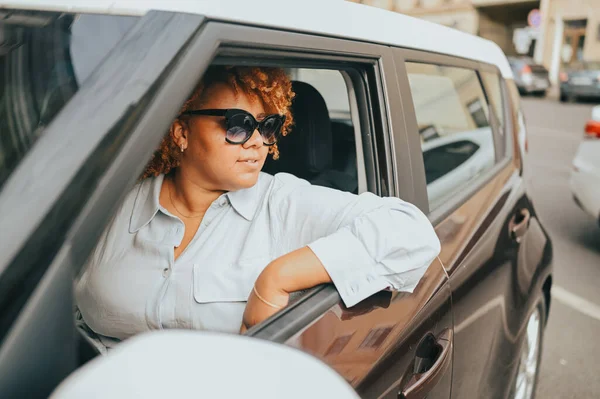 Happy young smiling African American woman red afro haired driver in sun glasses sitting in new brown car, smiling looking at camera enjoying journey. Driving courses and life insurance concept