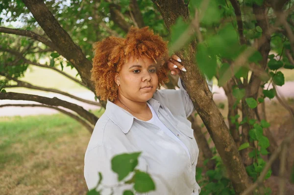 Retrato de moda de jovem naturalmente bonito plus size mulher afro-americana em terno calças leves com cabelo vermelho afro posando na natureza parque em folhagem verde. Diversidade e corpo conceito positivo. — Fotografia de Stock