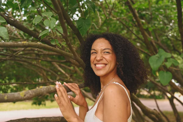 Fashion close up portrait of laughing atrakted young natural beautiful African American woman with afro hair and perfect toys approves posing in nature parkland in green foliage. — Stok Foto