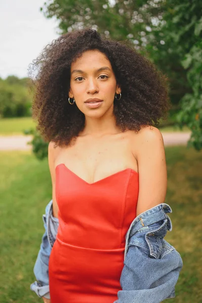 Potret mode menarik wanita muda Afrika Amerika yang cantik secara alami dengan rambut hitam afro dalam gaun merah dan jaket jins berpose di taman alam dalam dedaunan hijau. — Stok Foto