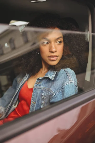 Cantik wanita muda Afrika Amerika berambut hitam driver dalam jaket jins duduk di mobil cokelat baru, menikmati perjalanan. Mengemudi kursus dan konsep asuransi jiwa — Stok Foto