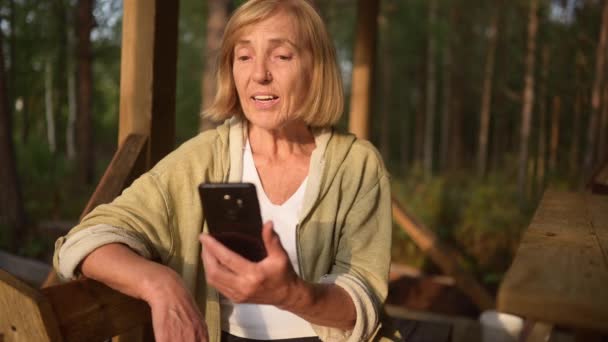 Attraktiv glad äldre kvinna som har videochatt utomhus, vinka. Samtal på mobiltelefon smartphone webbkamera. Positiv medelålders kvinna som gör videosamtal sittande på terrassen på landsbygden. — Stockvideo