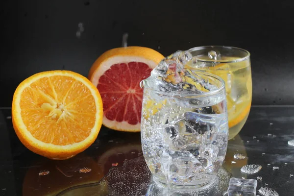 splashes of water over a glass of water and ice. Nearby are fruits orange and grapefruit, ice. On a black background. Refreshing cold ice drink.