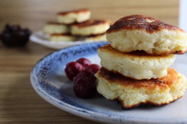 Üç krep peynirli kekler kiraz reçeli soslu bir masada bir tabağın üzerinde üst üste yığılmış slaytlar halinde dururlar. Yandan bak. Kahvaltı Rus Ukraynası. Tabaklar ve süzme peynir. Metin ve metin boşluğu için yer.