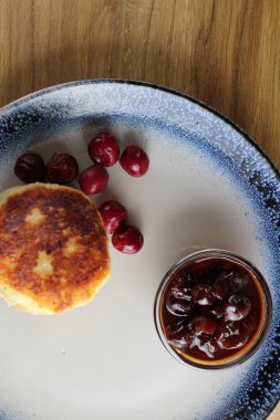 Üç krep peynirli kekler kiraz reçeli soslu bir masada bir tabağın üzerinde üst üste yığılmış slaytlar halinde dururlar. Yandan bak. Kahvaltı Rus Ukraynası. Tabaklar ve süzme peynir. Metin ve metin boşluğu için yer.