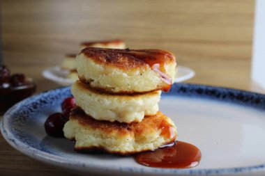 Üç krep peynirli kekler kiraz reçeli soslu bir masada bir tabağın üzerinde üst üste yığılmış slaytlar halinde dururlar. Yandan bak. Kahvaltı Rus Ukraynası. Tabaklar ve süzme peynir. Metin ve metin boşluğu için yer.