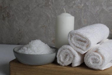 Spa tedavileri gevşetir. Evde vücut bakımı. Beyaz havlu mumları, banyo için tuz, gri bir arka planda, ahşap bir tepside yatar..
