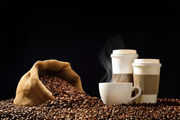 Cups of coffee with smoke and coffee beans in burlap sack on black background — Stock Photo, Image