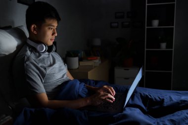 Genç adam geceleri yatağında dizüstü bilgisayar kullanıyor.