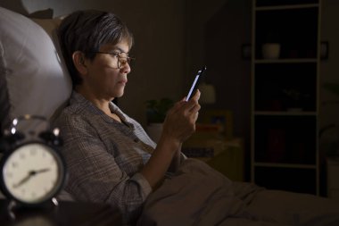 Gece yatakta yatarken akıllı telefon kullanırken gözleri yorgun ve ağrılı Asyalı bir kadın.