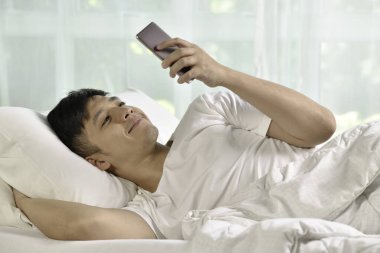 Genç Asyalı adam sabah uyandıktan sonra yatakta akıllı telefon kullanıyor.