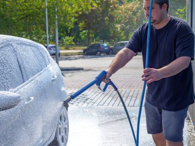 Elinde yüksek güçlü hortumlu bir adam arabasını oto yıkamada yıkıyor ve sürücü koltuğundan manzarayı seyrediyor. Kişisel temastan kaçının ve yalnız yıkayın..