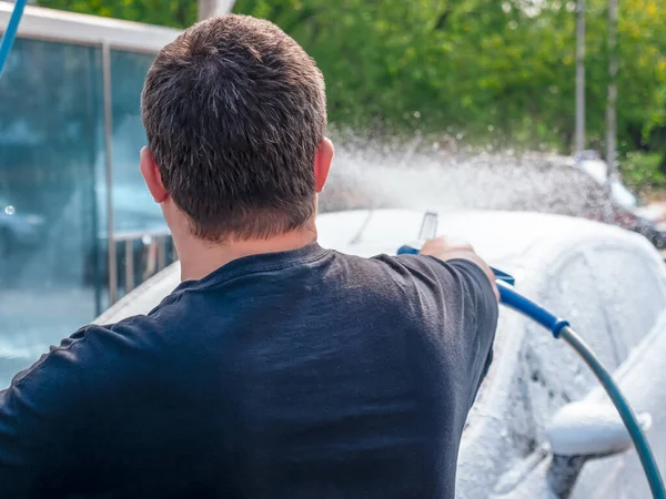 Yüksek güçlü hortumlu bir erkek eli, arabasını oto yıkamada yıkıyor ve sabunla kaplanmış bir perdeye bakıyor. Kişisel temastan kaçının ve yalnız yıkayın..