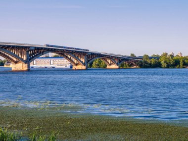 Dinyeper Nehri 'nin sağ kıyısından Kyiv şehrindeki ilk metro köprüsünün üzerinden mavi metro treni ve otelin arkasında olduğu yere bakın. Mavi gökyüzüyle güneşli bir gün.