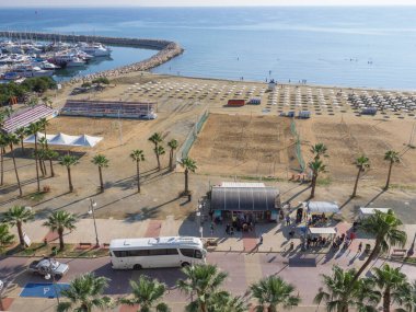 Merkez otobüs durağına tepeden bakan üst hava manzarasında Finikoudes Palmiye ağacı gezinti alanı, Akdeniz, liman ve plaj voleybol sahasında güneşli bir günde Kıbrıs Rum Kesimi 'nin eski Larnaka kentinde bekleyen insanlar görülüyor..