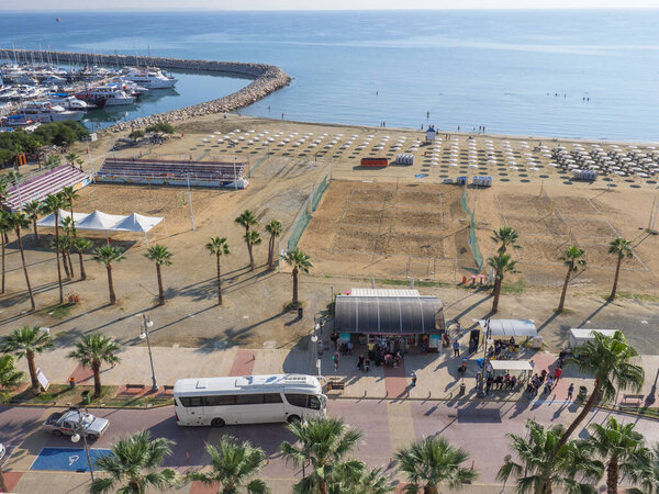Top aerial view overlooking the central bus stop with people waiting at Finikoudes Palm tree promenade, mediterranean sea, yachts in the port and beach volleyball court on a sunny day in Larnaca old town, Cyprus.