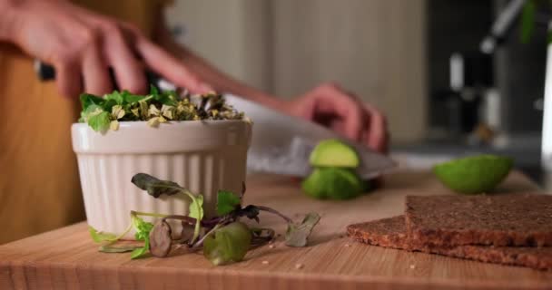 Selektiver Fokus auf Mikrogemüse in einer Schüssel, Frau schneidet Avocado im Hintergrund. — Stockvideo