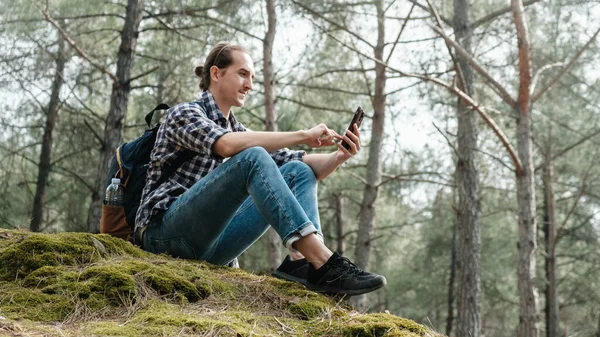 リュックを背負ったハンサムな若い男がスマートフォンを手に森の中の岩の上に座っている 観光客はスマートフォンの地図やナビゲーションを見ています 観光客はスマートフォンでインターネットを使用します — ストック写真