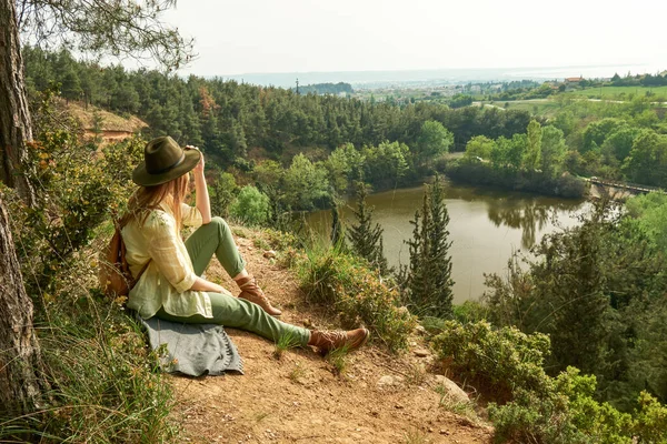 山の頂上にはリュックと帽子を持つ若い女性が座っており 川との美しい風景を楽しんでいます — ストック写真