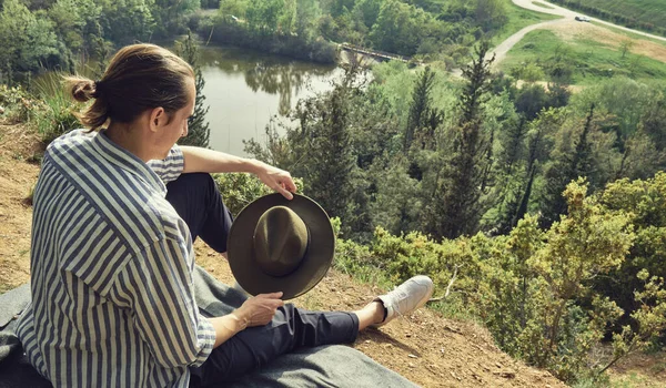 山の頂上にはリュックと帽子を持つ男の旅行者が座っており 川との美しい風景を楽しんでいます — ストック写真
