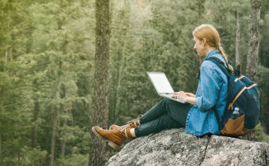 Sırt çantalı genç bir kadın ormanda oturuyor ve dizüstü bilgisayarla çalışıyor. Çalışma konsepti. Serbest çalışan biri.