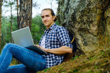 Adam ormanda oturuyor ve dizüstü bilgisayarla çalışıyor.
