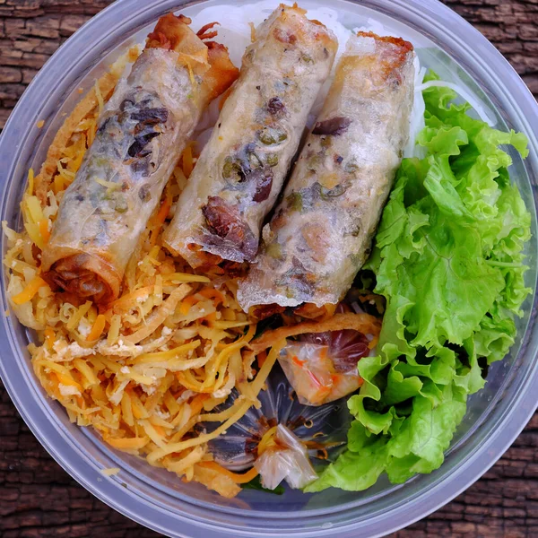 Top view serving from order online in plastic bowl, vegan rice noodles with fried spring rolls and salad, sauce, veg food for Vietnamese vegetarian on wooden background