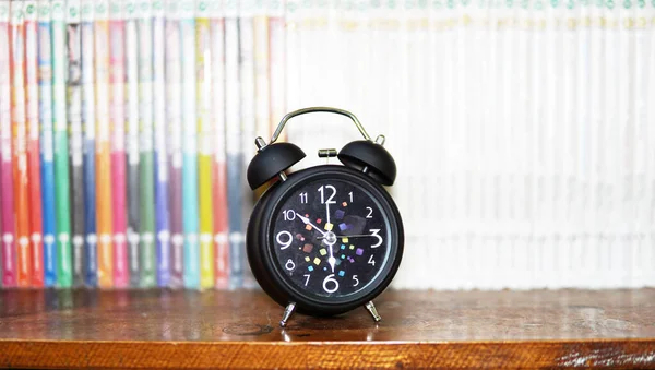 Black table clock on wooden bookshelf.