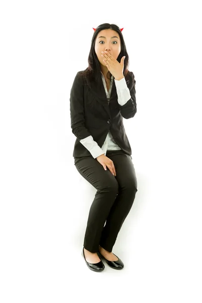 Surprised attractive Asian young businesswoman sitting on stool in devil horns covering her mouth by the hands isolated over white background — Stok fotoğraf