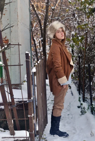 Foto de retrato de una hermosa chica con abrigo y sombrero de piel de pie cerca de la cerca y la pared de la casa en el fondo de la naturaleza de invierno — Foto de Stock