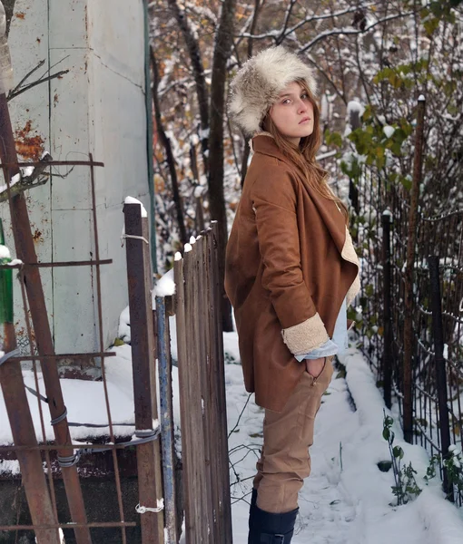 Foto de retrato de una hermosa chica con abrigo y sombrero de piel de pie cerca de la cerca y la pared de la casa en el fondo de la naturaleza de invierno — Foto de Stock