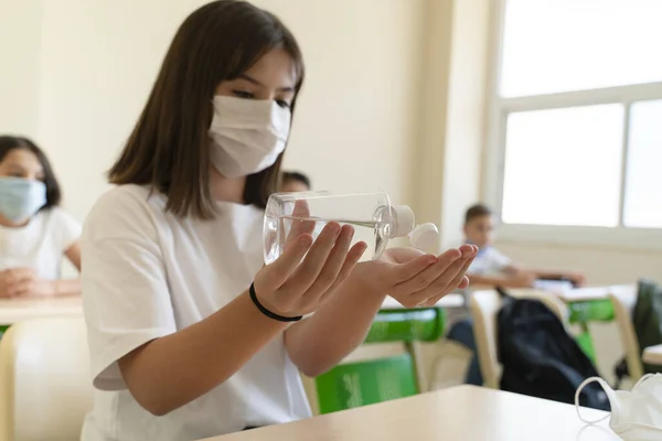 Schoolgirl with face mask cleaning hands with disinfecting antibacterial gel in the classroom.