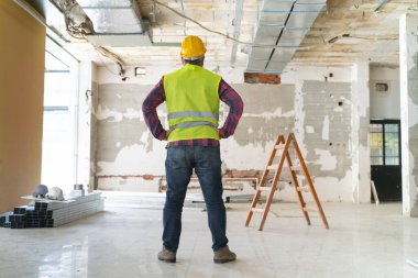 İnşaat alanında çalışan, koruyucu kasklı, inşaat aletleriyle ayakta duran bir inşaat işçisi.