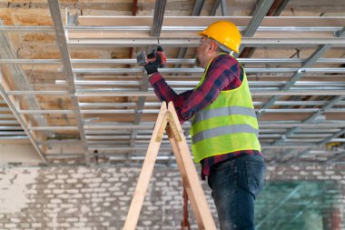 Bir merdivende tamirci elinde bir makineyle tavana sondaj yapıyor.